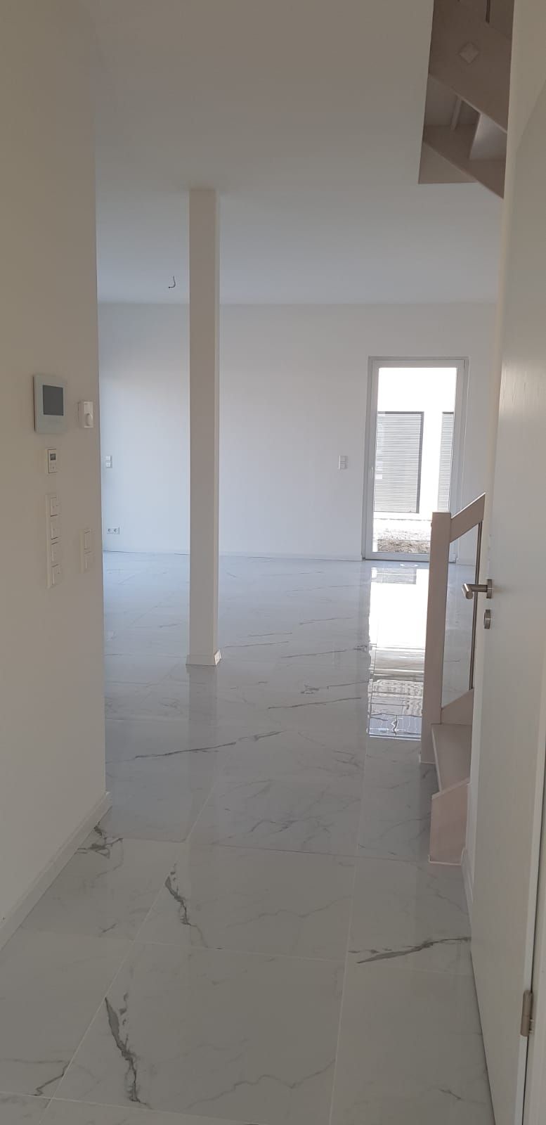 Innenansicht eines modernen, leeren Hauses mit weißen Wänden, Böden und einer Tür, die zu einem hellen Außenbereich führt.