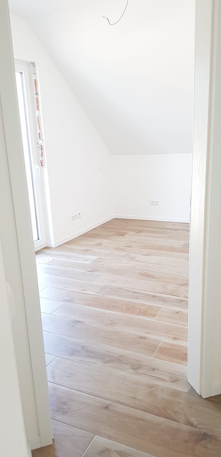 Blick durch eine Türöffnung in einen weiß getünchten Raum mit Holzboden. Der Raum hat eine Dachschräge und ein Fenster.