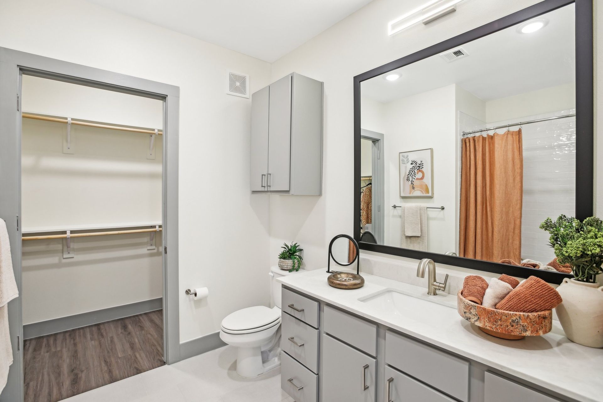Bathroom with gray cabinets, white countertop, large mirror, walk-in closet, and orange shower curtain.