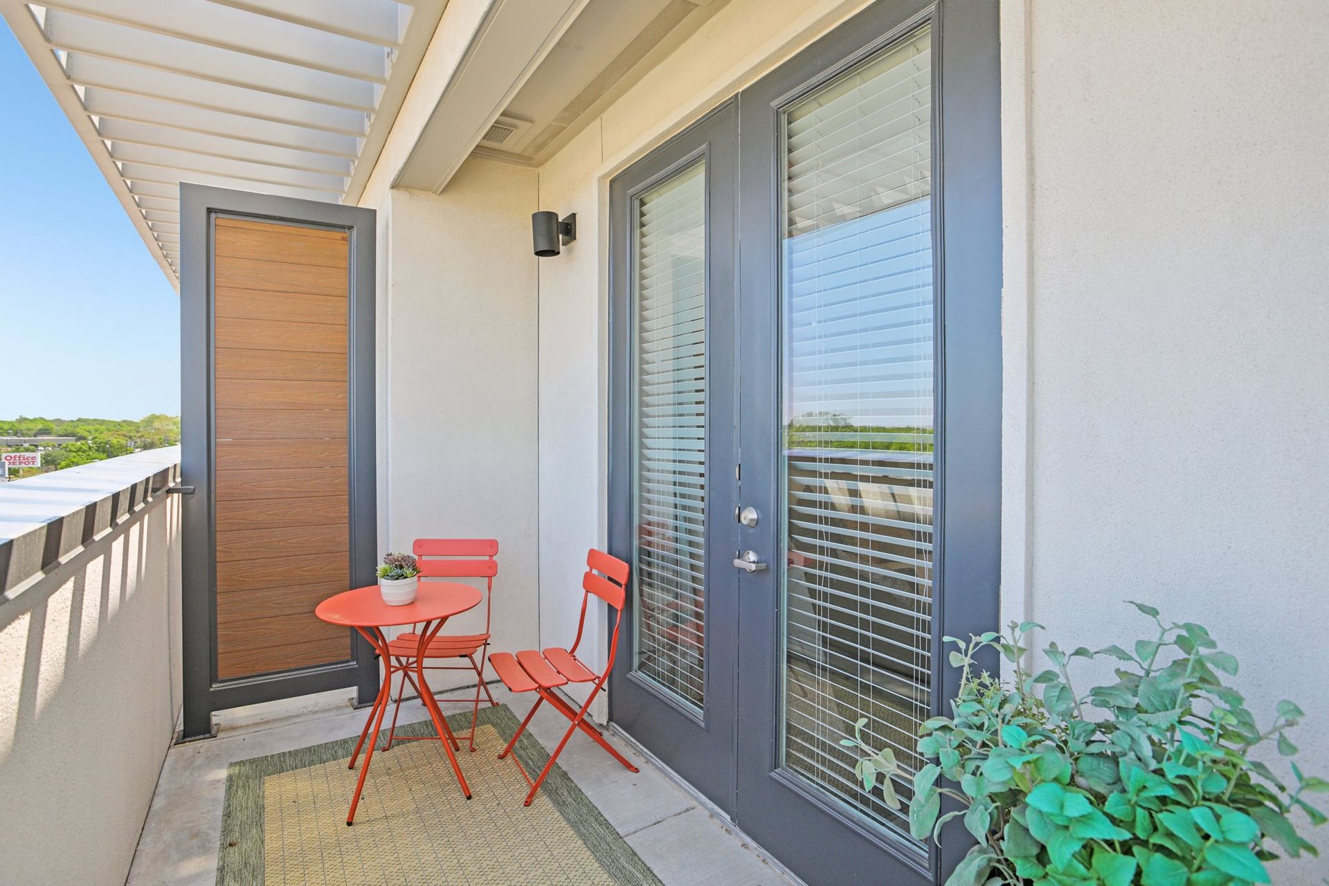 Small balcony with red table and chairs, green plant, and grey doors.