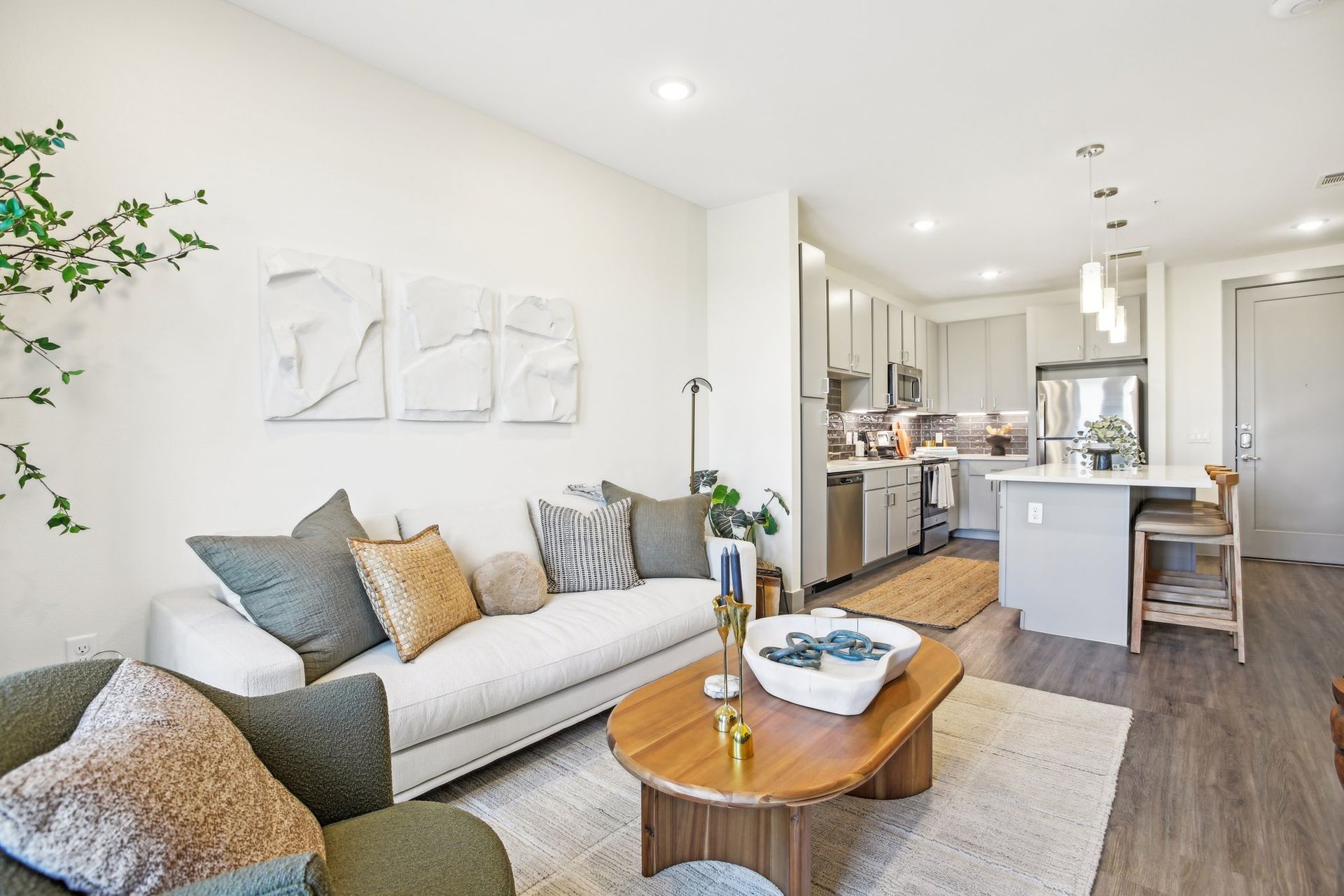 Living room with cream walls, open to a kitchen. Features a sofa, arm chair, rug, coffee table, and artwork.