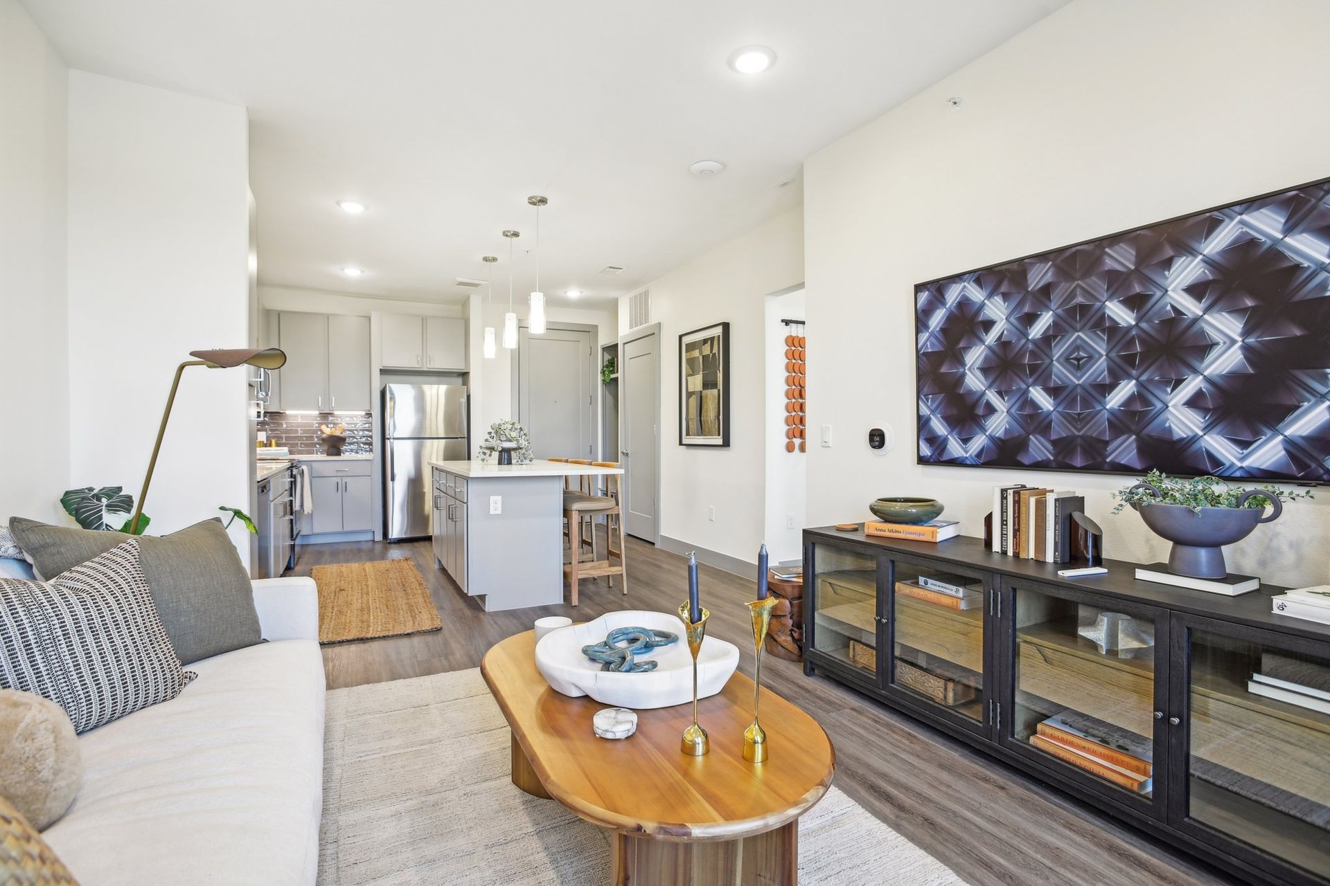 Living room with open kitchen, white walls, wooden furniture, and abstract TV art.
