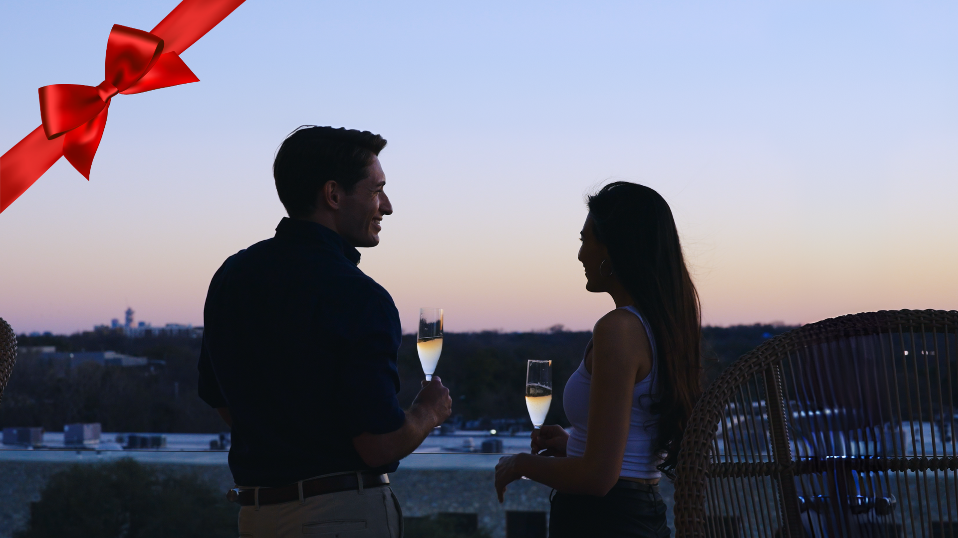 Couple toasting with champagne on a rooftop at sunset. Red bow overlay.