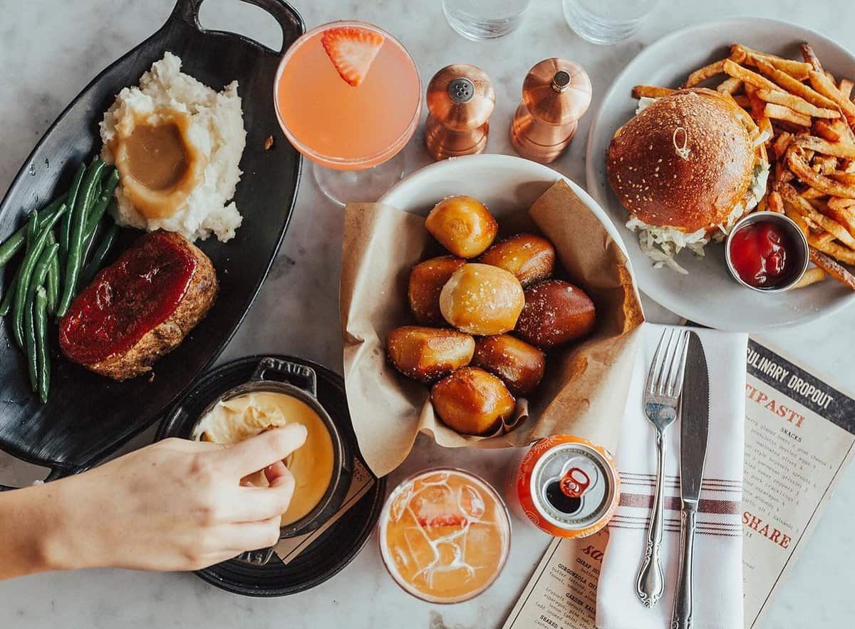 Overhead shot of a table with various dishes: steak, mashed potatoes, burger, fries, pretzels, drinks, and a hand dipping pretzel into cheese.