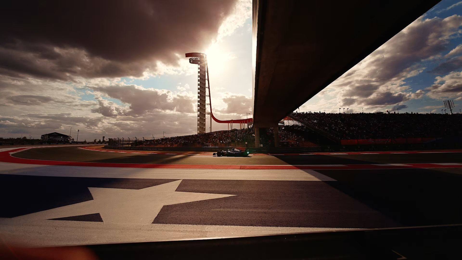 Race car speeding on track, under a bridge, with a cloudy sky and grandstands.
