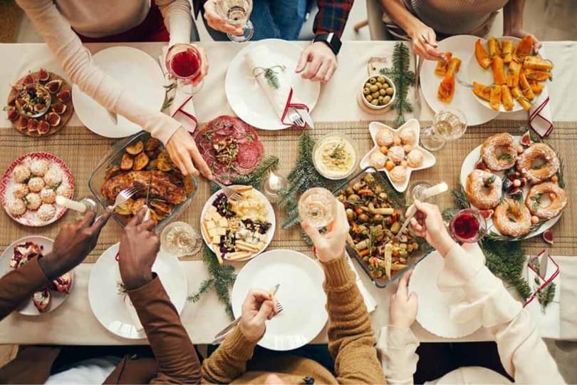 People reaching for food at a festive table setting with diverse dishes, drinks, and hands raised in a toast.