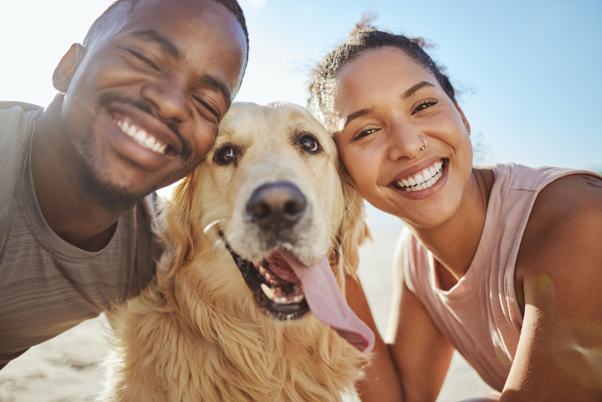 Couple smiling with a golden retriever; they are outdoors in the sun.