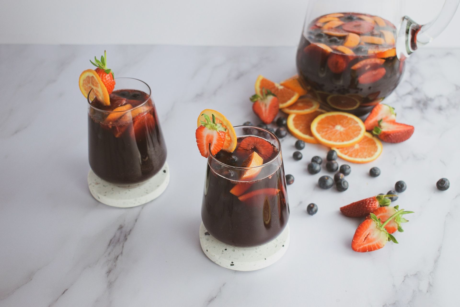 Sangria with fruit in two glasses and a pitcher on a marble surface.