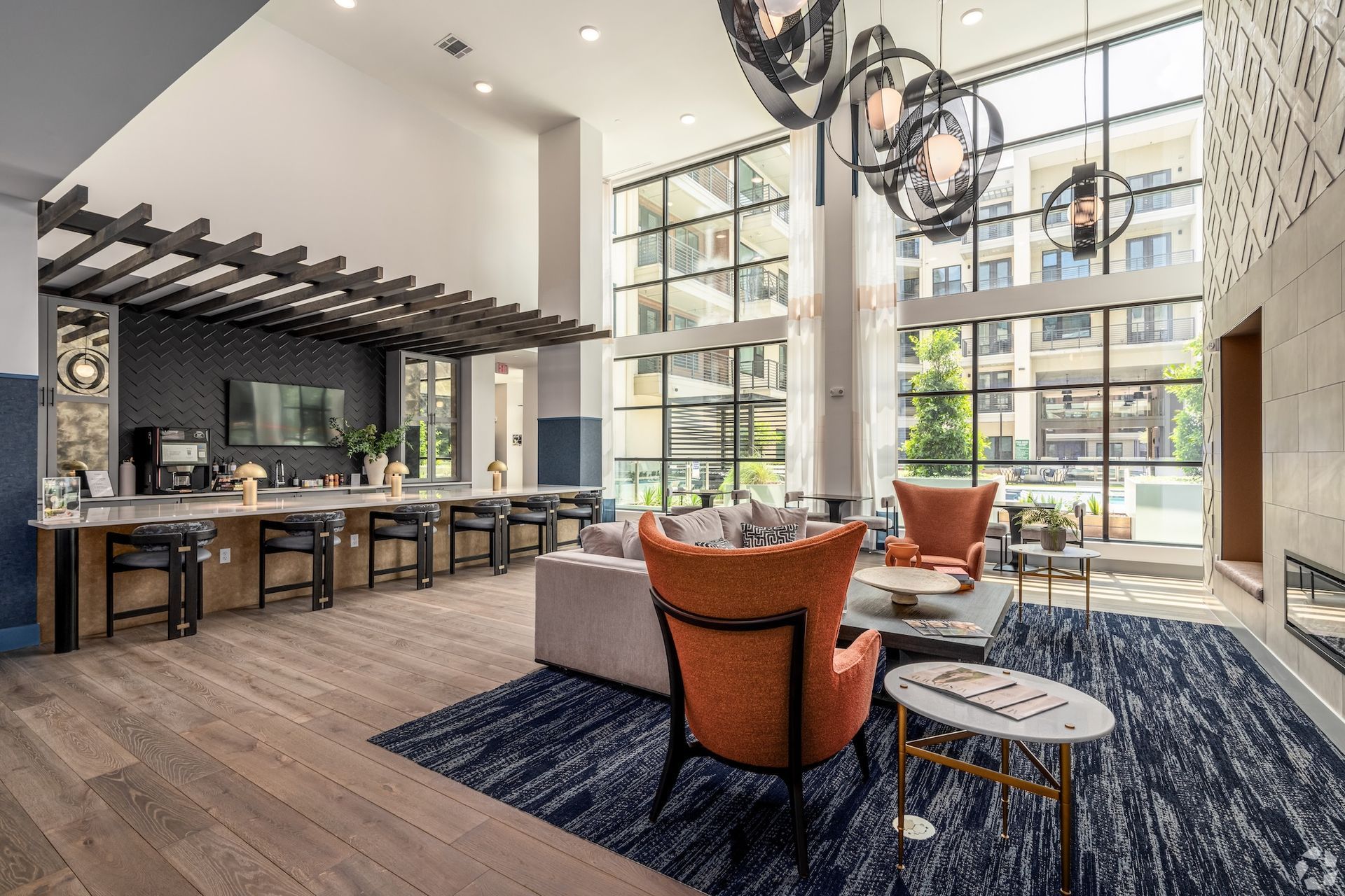 Lobby with seating, kitchen, and large windows. Orange chairs, blue rug, wooden floor.
