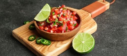 A bowl of salsa with limes and jalapenos on a wooden cutting board.
