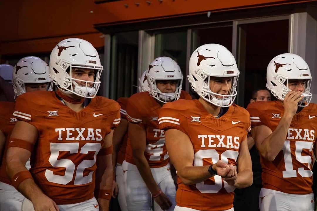 Texas Longhorns football players in orange jerseys and white helmets enter a stadium.