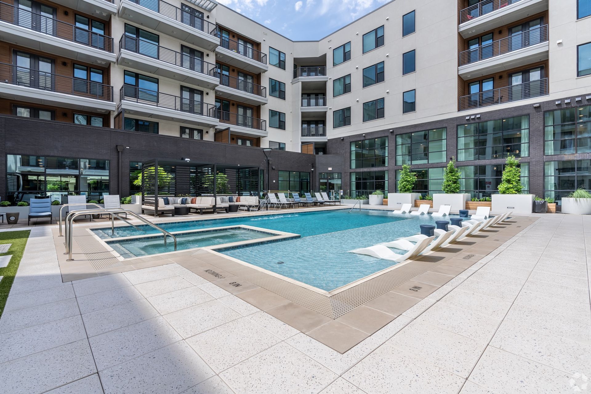 Swimming pool in courtyard of modern apartment complex. Lounge chairs, hot tub, and glass walls visible.