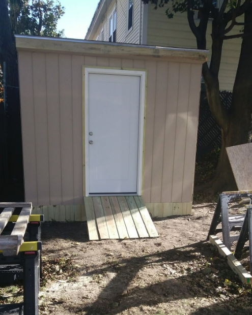 Small beige shed with a white door and short wooden ramp in a yard