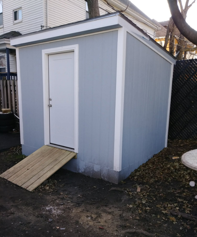 Small blue-gray shed with white trim and door, plus a wooden ramp in a backyard.