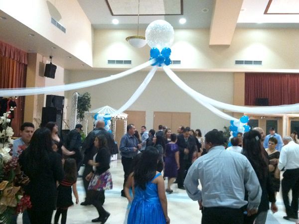 Guests mingle on a decorated dance floor with white and blue drapes and balloons in a banquet hall.