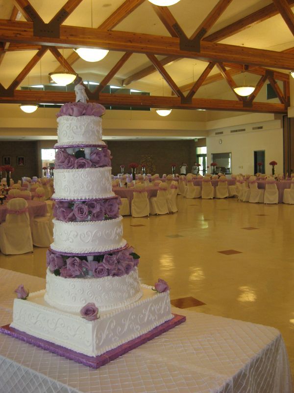 Tall white and lavender tiered cake on a table in a banquet hall with round tables in the background