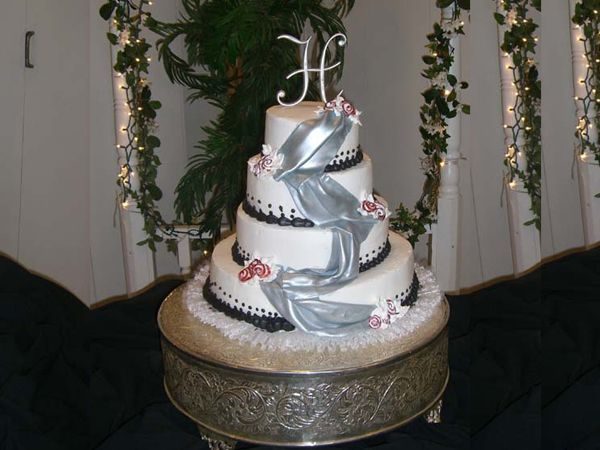 Three-tier silver-and-white wedding cake with roses on an ornate stand, against greenery backdrop