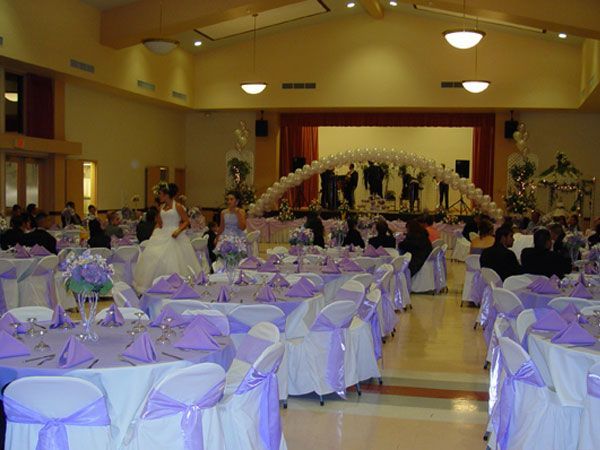 Banquet hall with round tables dressed in white and lavender, guests seated beneath a lit arch at the front.