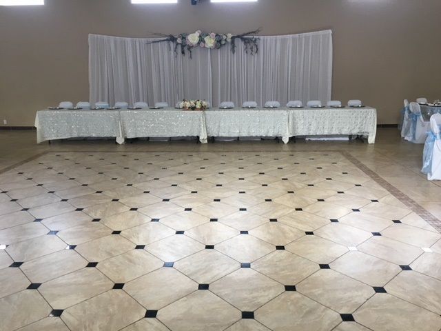 Empty banquet hall with tiled floor, long white-clothed head table, and rows of white chairs along the back wall