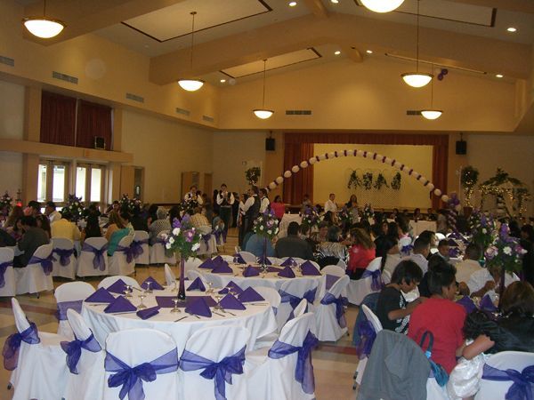 Banquet hall with round tables set in purple and white, guests seated, and a decorated stage at the back