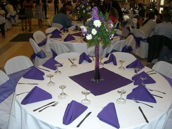 Round banquet table with white chairs and purple napkins, set with glasses and a purple floral centerpiece.