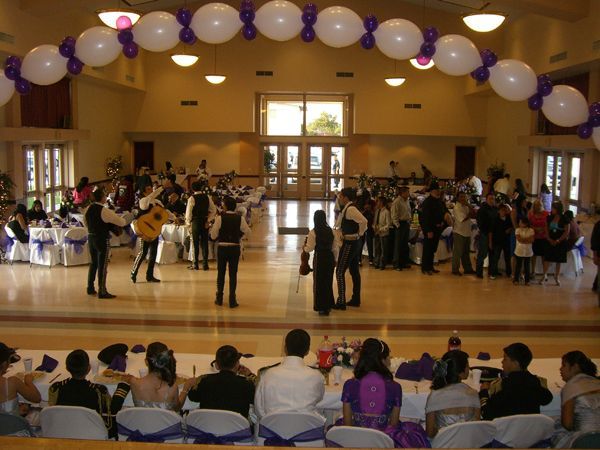 Students dancing on a gym stage under purple and white balloons, with an audience seated in front.