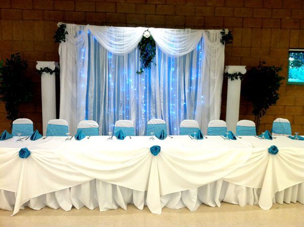 Decorated banquet table with white drapes, blue accents, and rows of chairs in a hall