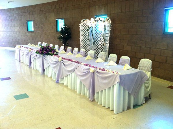 Banquet table with white drapes and flower arrangements along a stone wall in a hall