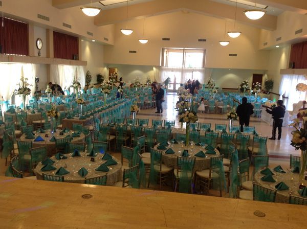 Decorated banquet hall with teal chairs and tables arranged for an event, with a few people standing near the center.