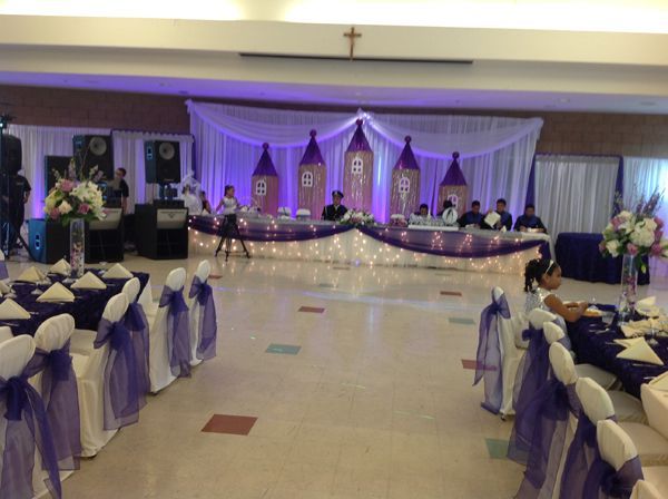 Decorated banquet hall with purple drapes, round tables, and a head table on a lit stage