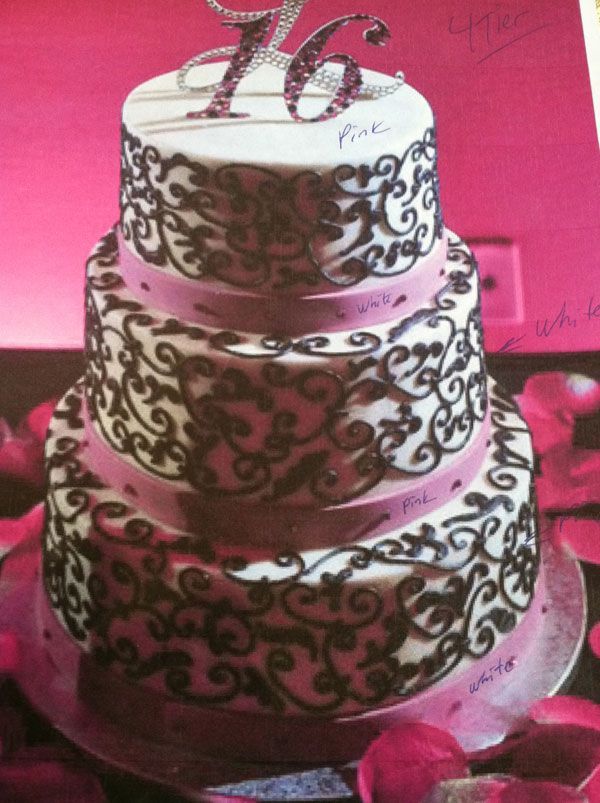 Three-tier white wedding cake with ornate dark icing scrollwork and pink ribbon accents on a pink background