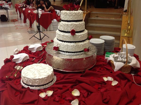 Multi-tiered white wedding cake on a red-draped table with rose petals and stacked plates nearby