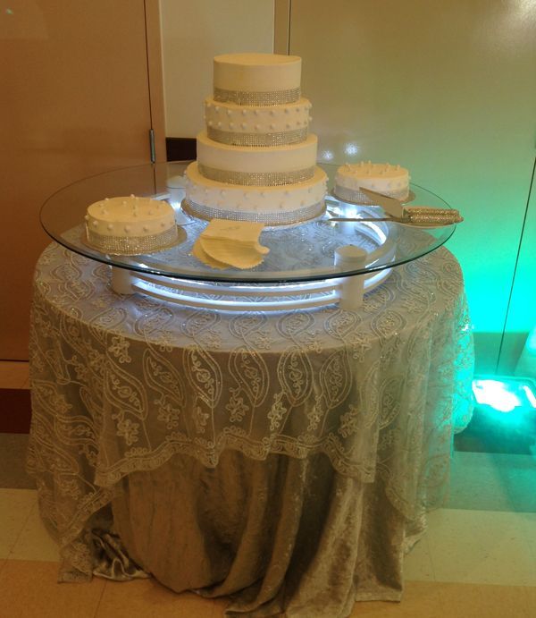 Tiered white wedding cake on a lace-covered table, with a smaller cake and serving plates on a glass top.