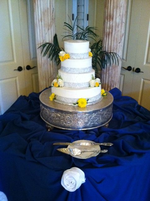White tiered cake with yellow flowers on a silver stand, set on a blue-draped table at an indoor event