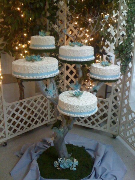 Tiered white wedding cake stand with several cakes, blue ribbon accents, and flowers in a garden-like display
