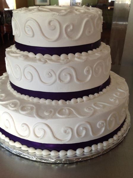 Three-tier white wedding cake with purple ribbon accents and silver scroll details