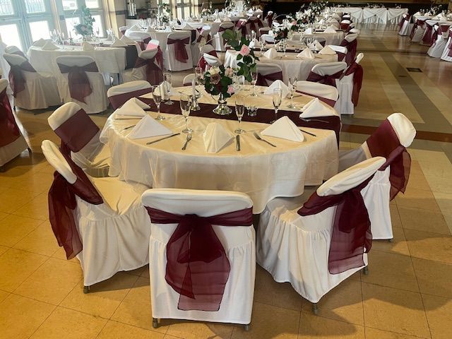 Round banquet tables set for an event, with white chair covers and burgundy sashes in a decorated hall