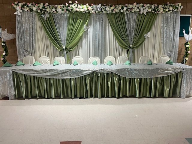 Decorated banquet table with green-and-white drapes, floral garland, and white chairs on a stage
