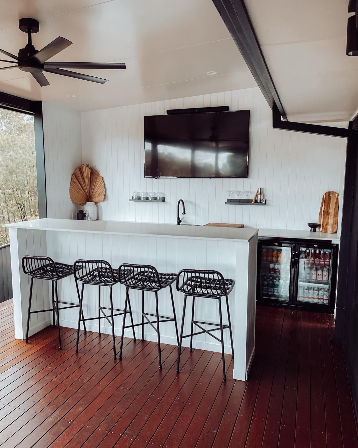 Outdoor bar with white counter, black stools, and mini-fridges. TV mounted on white wall.