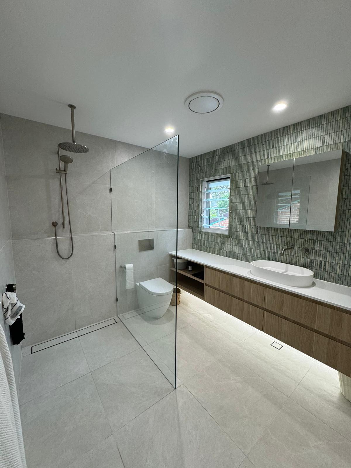 Modern bathroom with gray tiled shower, toilet, floating wood vanity, and decorative green mosaic tile accent wall.