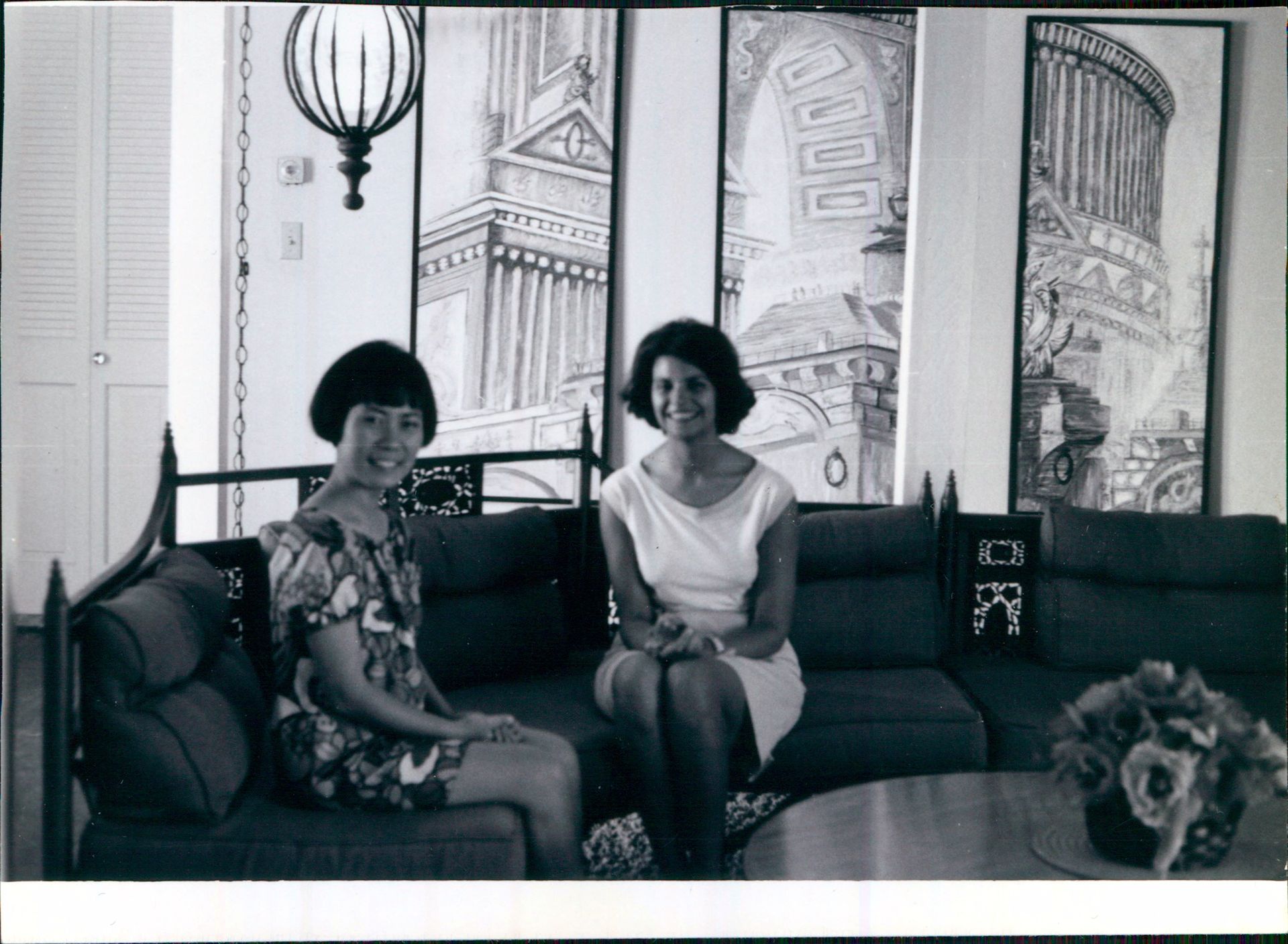 Two women sit on a couch in front of painted architectural panels. One wears a floral dress, the other a white dress.