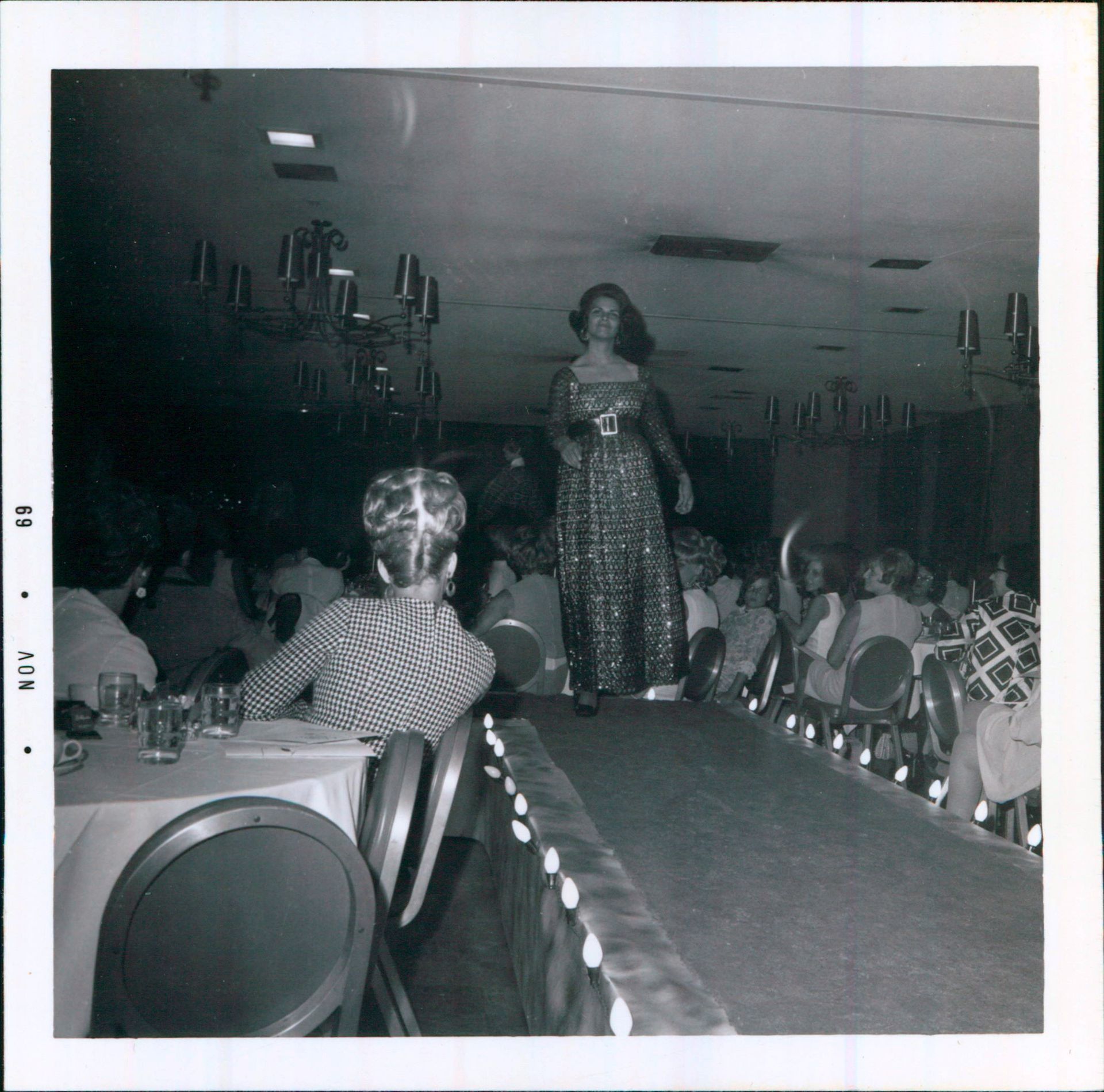A model in a long, patterned dress walks a runway during a fashion show in a banquet hall.