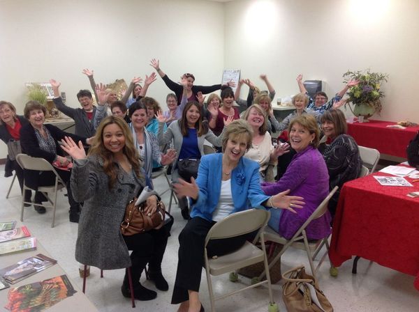 Group of smiling women in a room, arms raised, celebrating. Tables with red cloths.