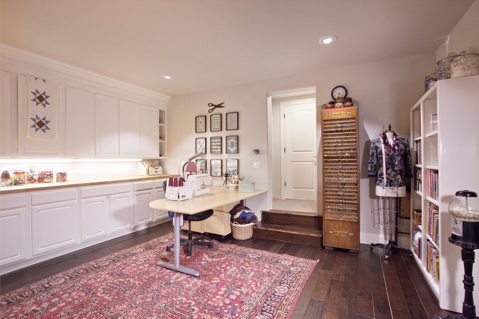 A craft room with white cabinets, a sewing machine, and a red patterned rug on a wood floor.