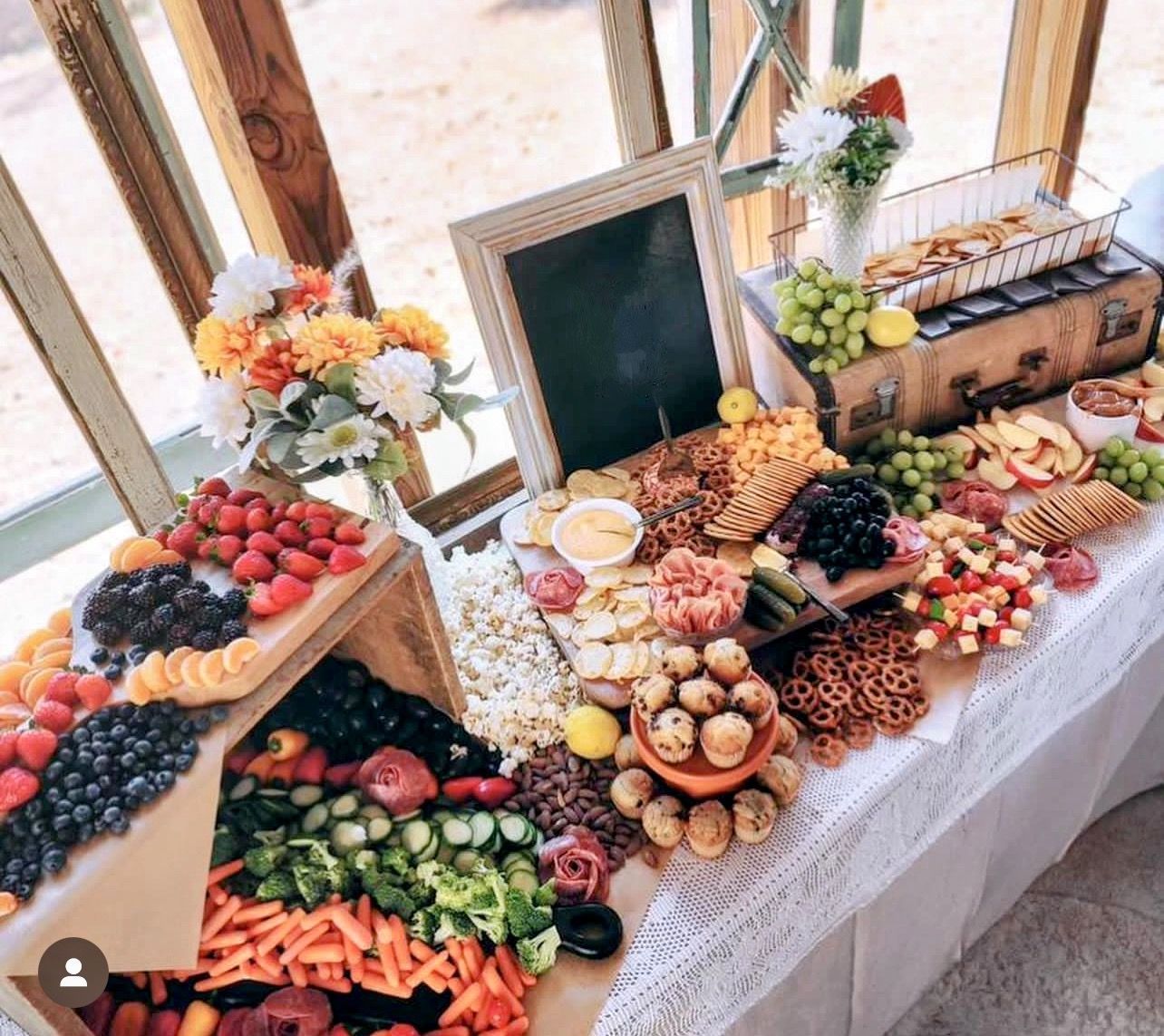 Charcuterie spread with fruits, vegetables, cheeses, and crackers, on a table with flowers and a chalkboard sign.