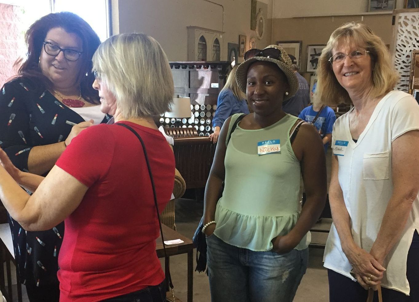Four women smiling inside a store; one in a red shirt, one in a light green tank top, and two wearing glasses.