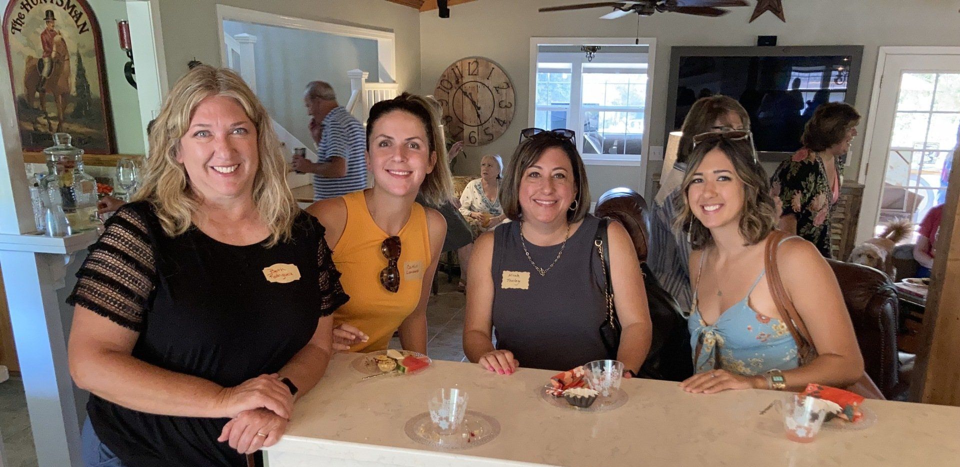 Four women smiling at a bar; others in background. Inside with food and drinks.
