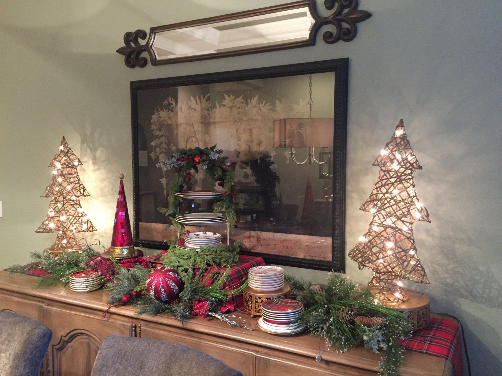 Christmas decorations on a buffet: lighted trees, garland, ornaments, red and green, mirror in background.