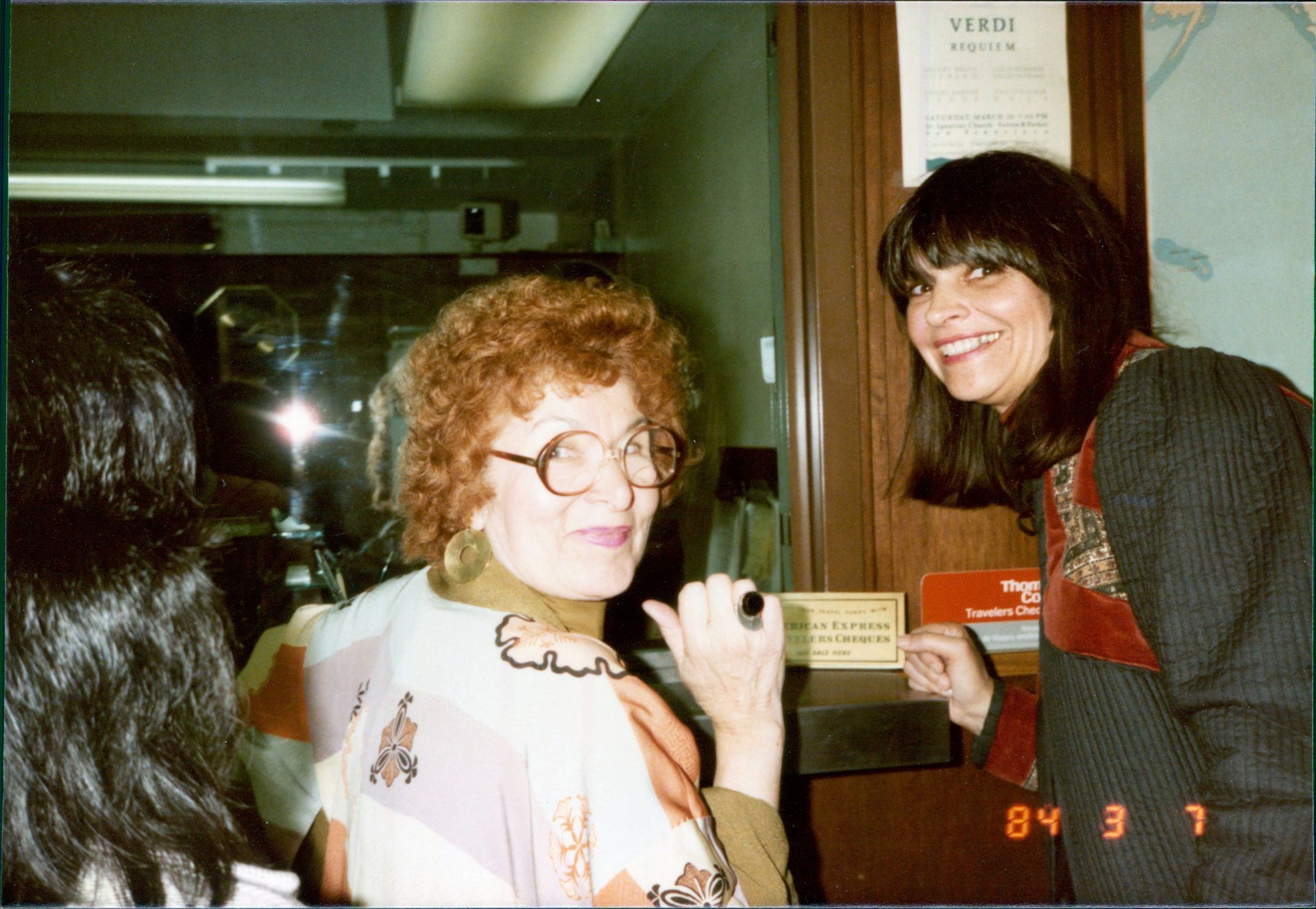 Two women smiling near a window; one with curly red hair, the other with long dark hair. March 1, 1984.