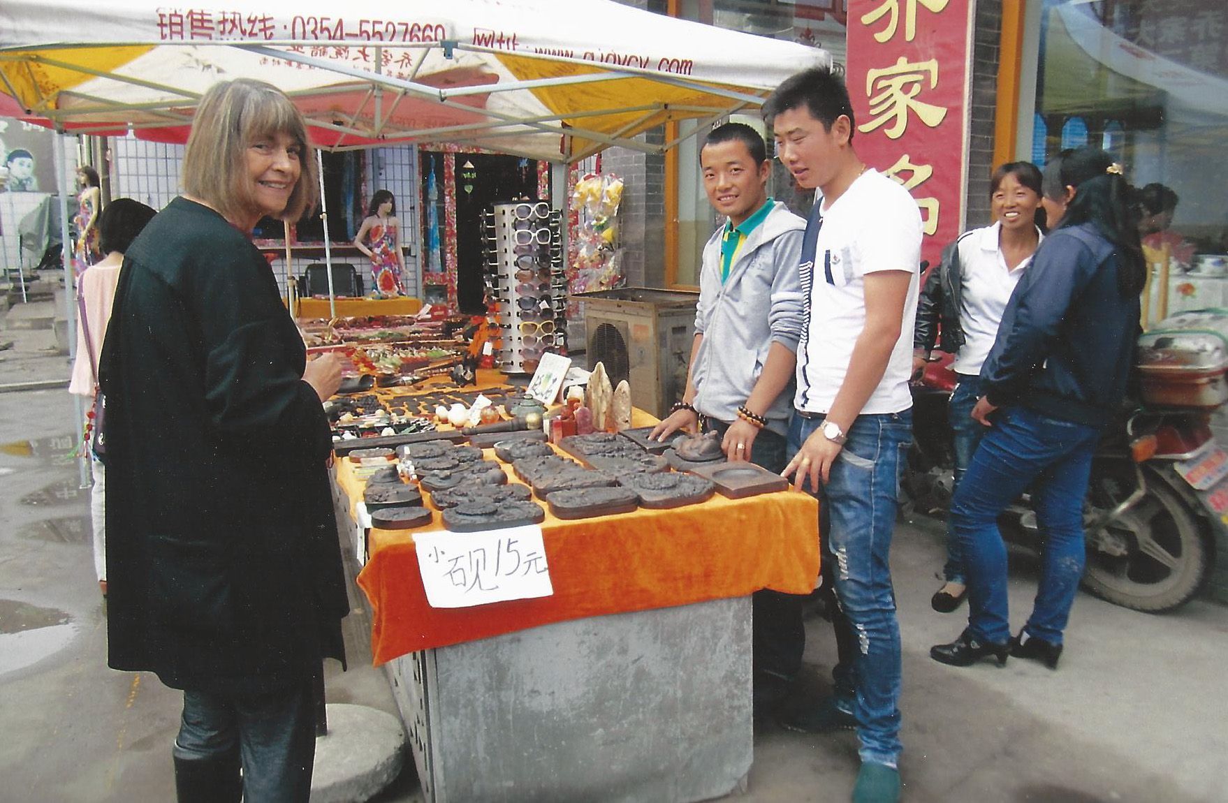 A woman shops at a market stall with other people, a man and a woman are looking at the products.